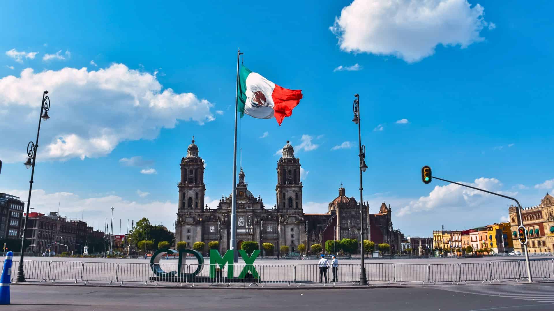 A large Mexican flag waves in front of the Mexico City Metropolitan Cathedral in the Zócalo square.
