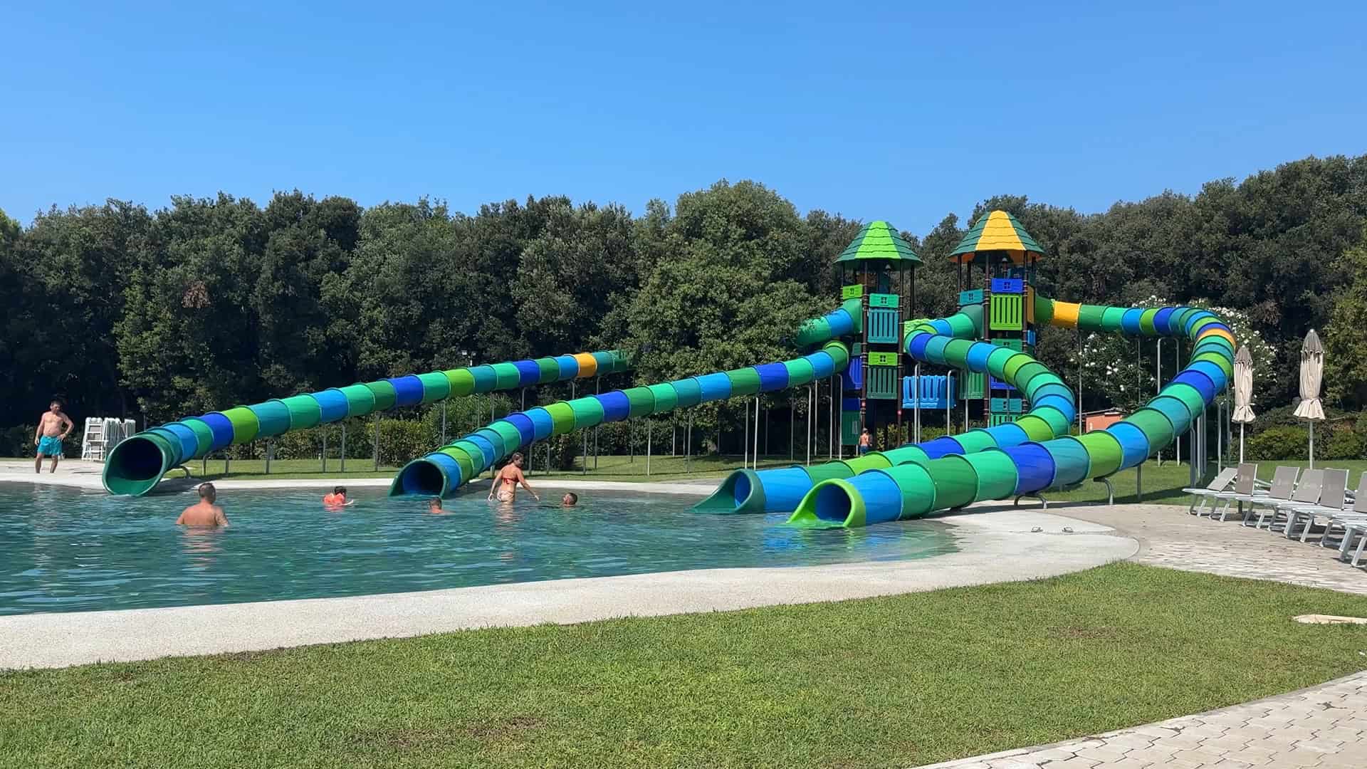 Large blue and green water slides at Parco della Gallinara lead into a pool where people are swimming on a sunny day.