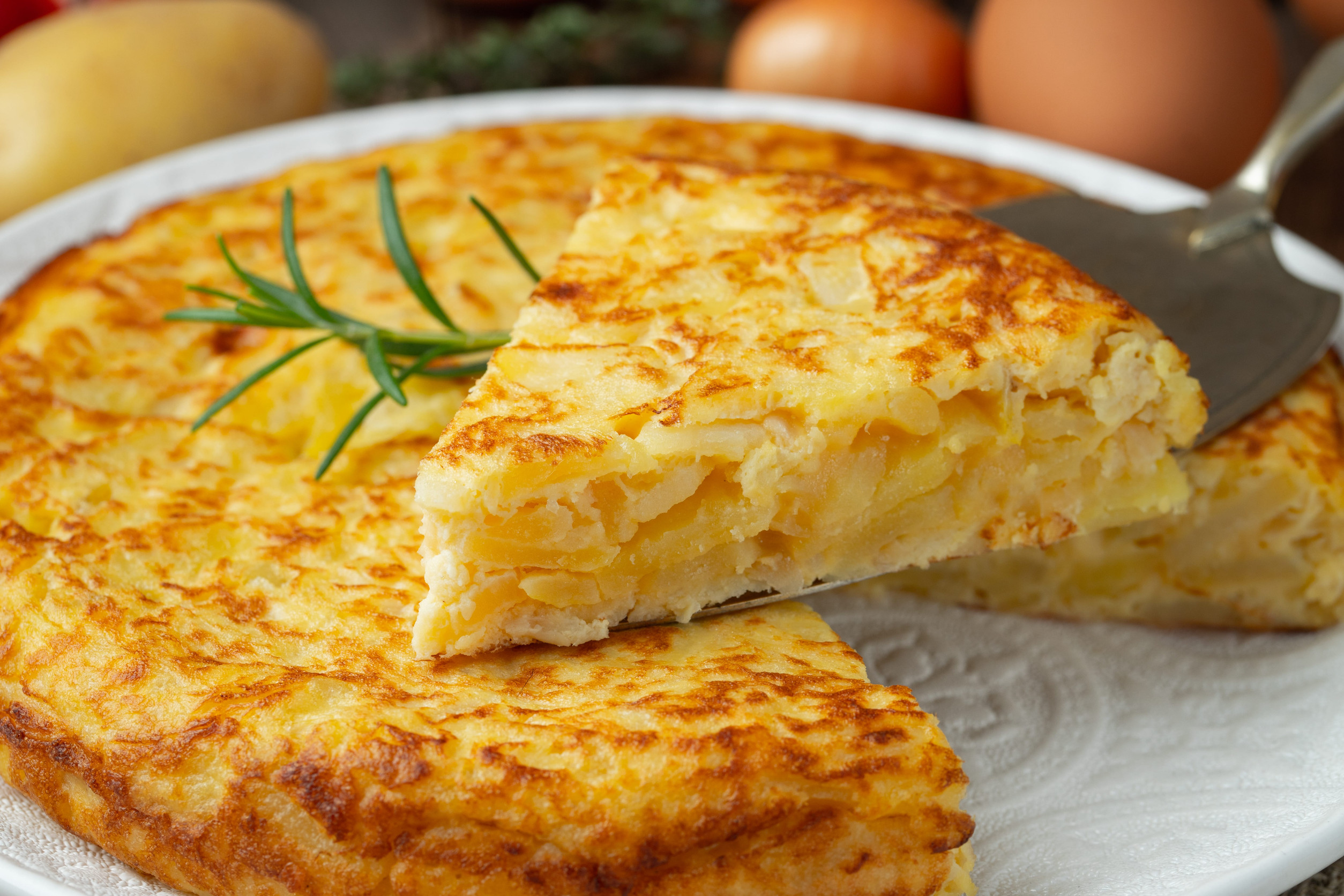 A slice of Spanish tortilla being served from a whole tortilla on a white plate, garnished with rosemary&mdash;an iconic vegetarian food and one of Spain&rsquo;s top 10 classic dishes.