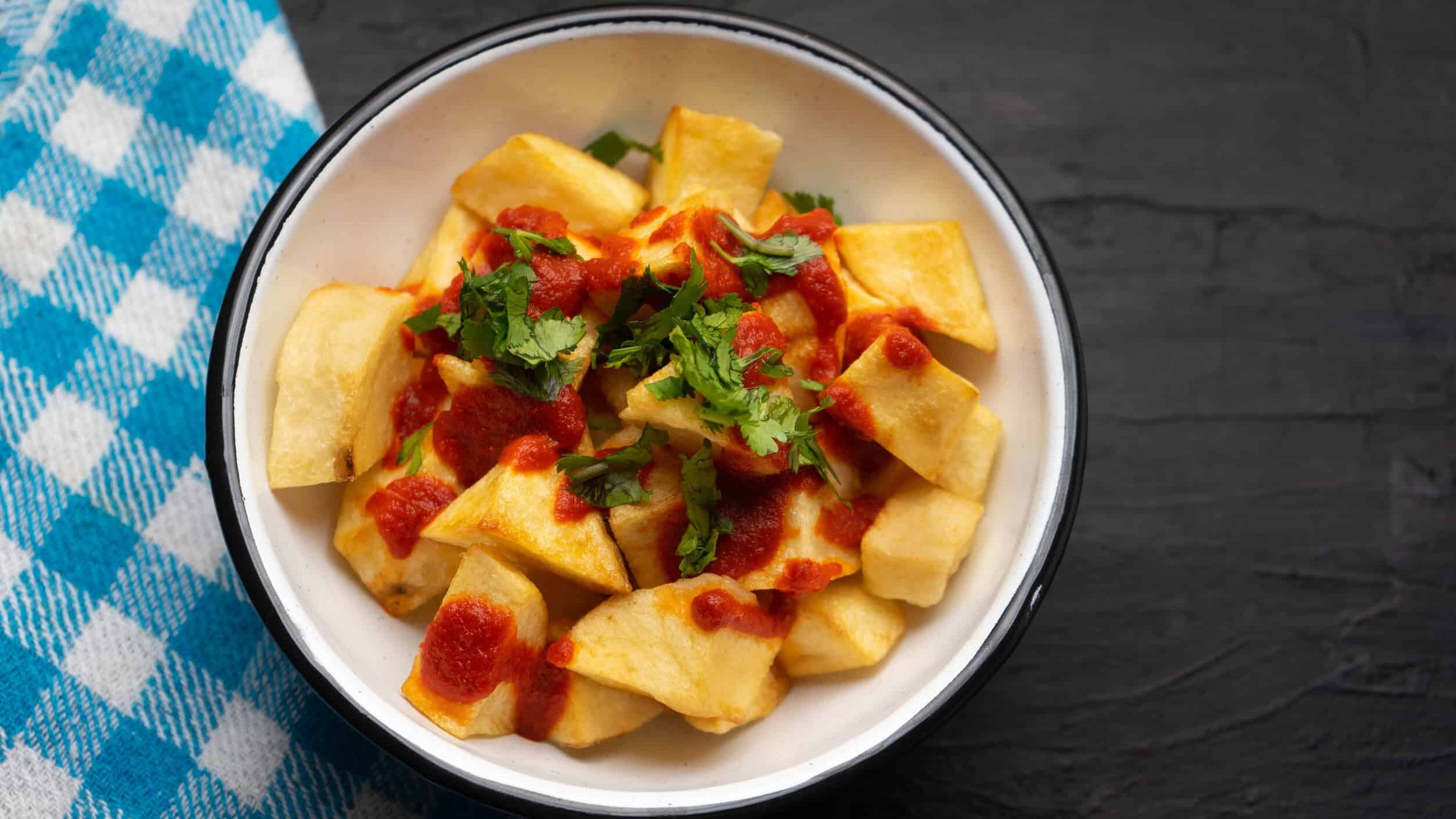 A bowl of patatas bravas topped with red sauce and chopped cilantro, next to a blue checkered napkin&mdash;a classic among vegetarian dishes Spain is famous for.