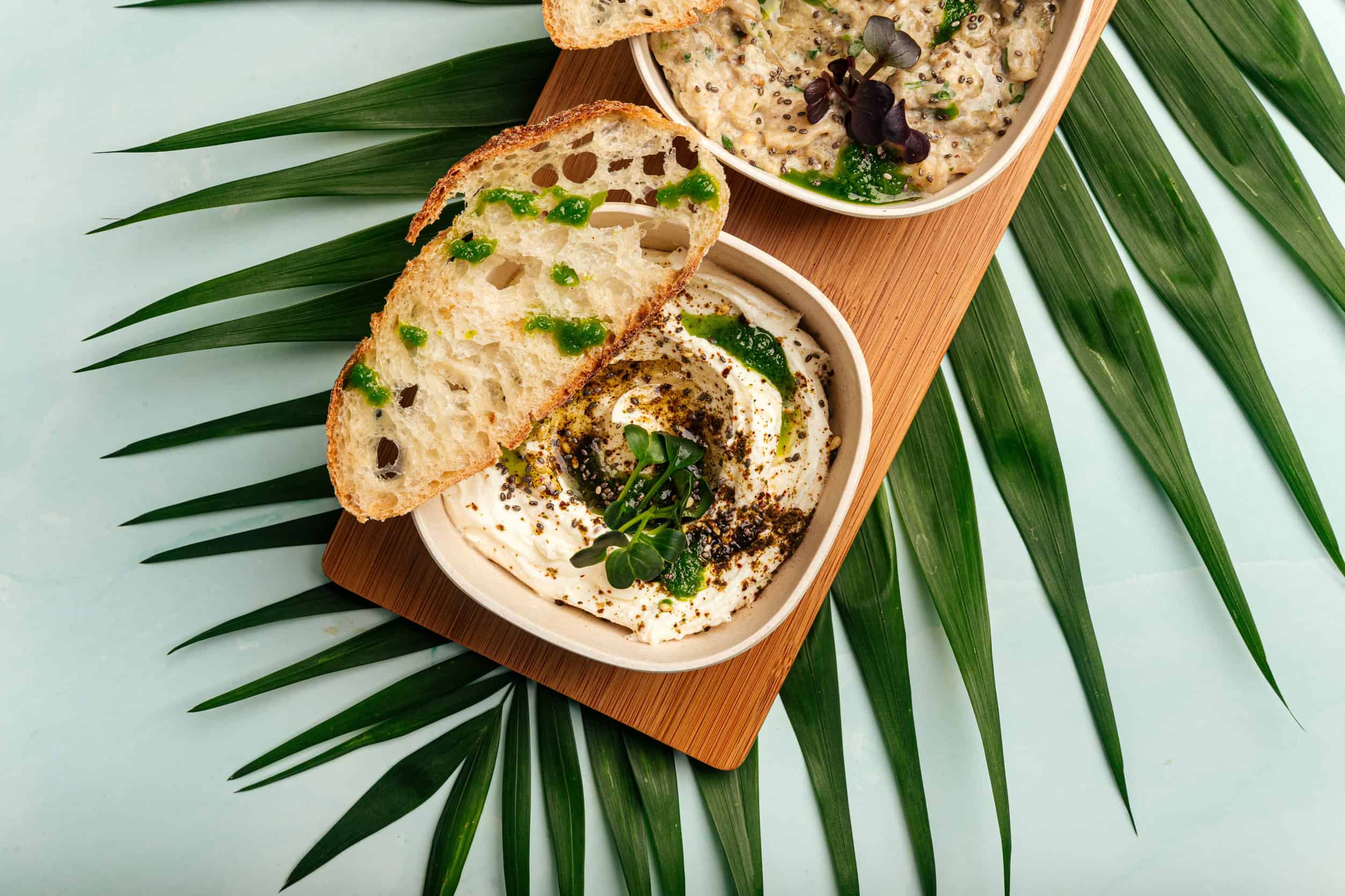 Two bowls of dip with herbs, served with artisan bread on a wooden board over green palm leaves—a beautiful example of Vegetarian Dishes in Tunisia.