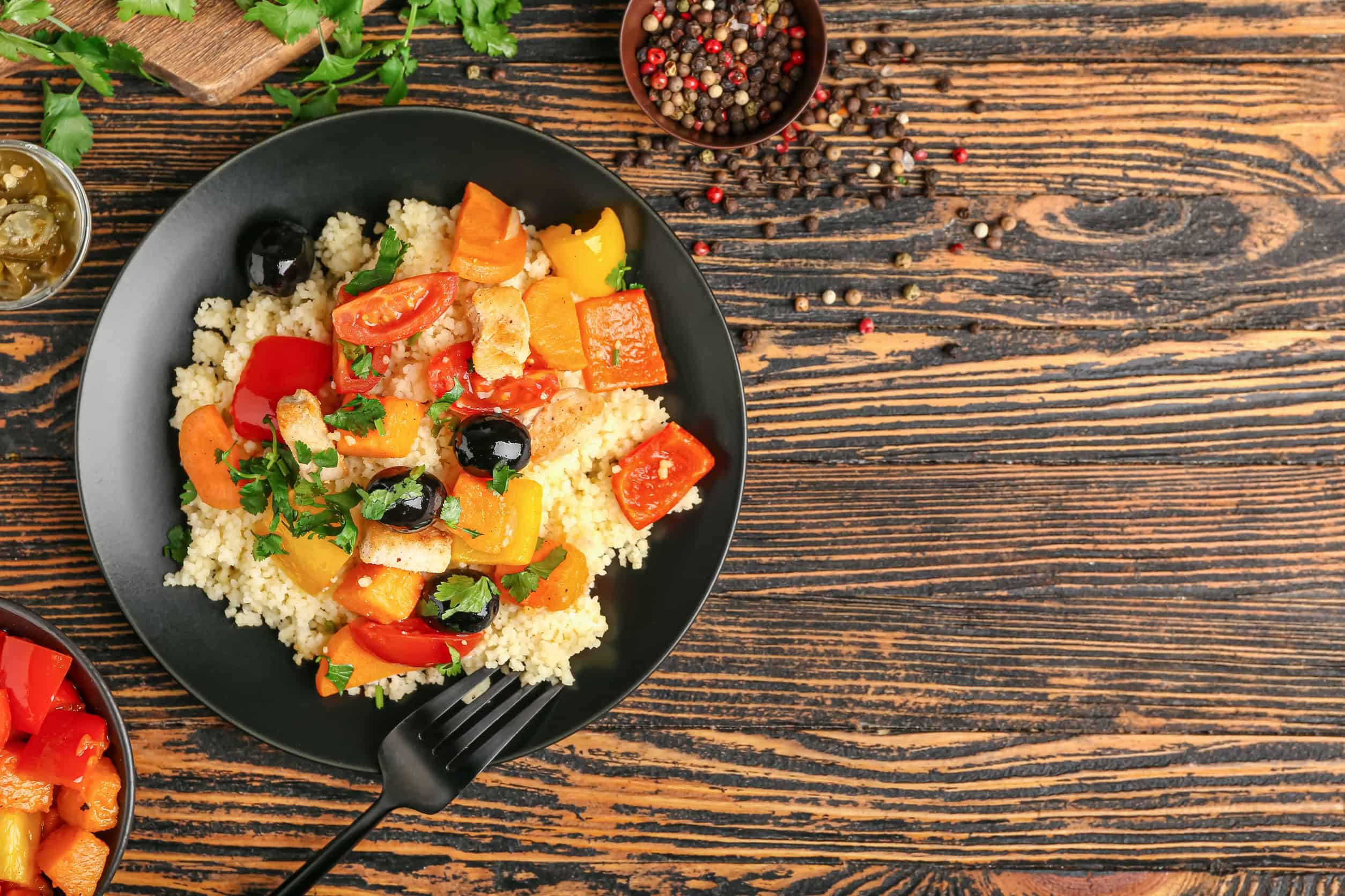 A black plate with couscous, roasted vegetables, olives, and herbs on a rustic wooden table showcases one of the flavorful vegetarian dishes in Tunisia.
