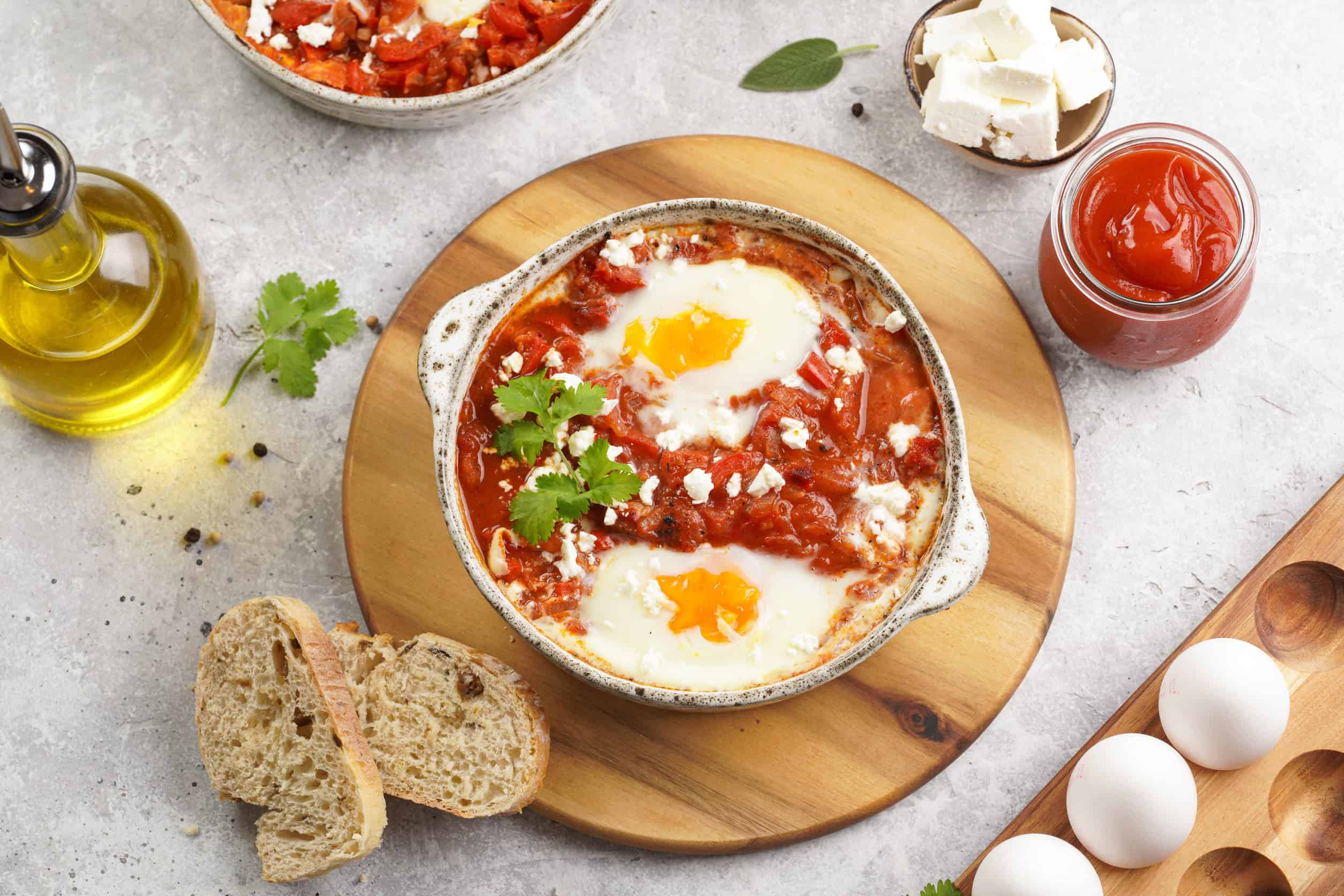 Bowl of shakshuka with eggs, bread slices, olive oil, feta cheese, tomato sauce, and herbs on a wooden board—a classic among vegetarian dishes in Tunisia.