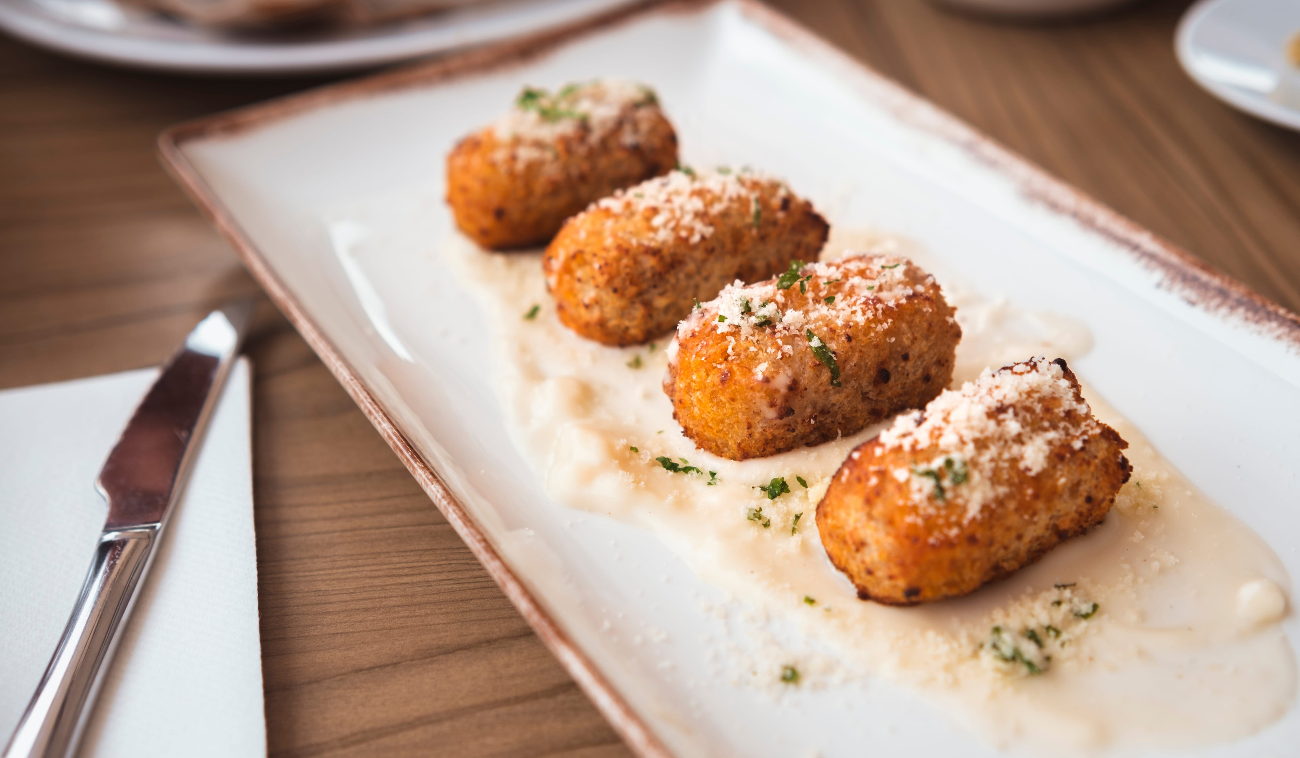 Four golden croquettes, a Top 10 pick for Vegetarian Food in Spain, topped with grated cheese and herbs, served on a rectangular plate with a creamy sauce.