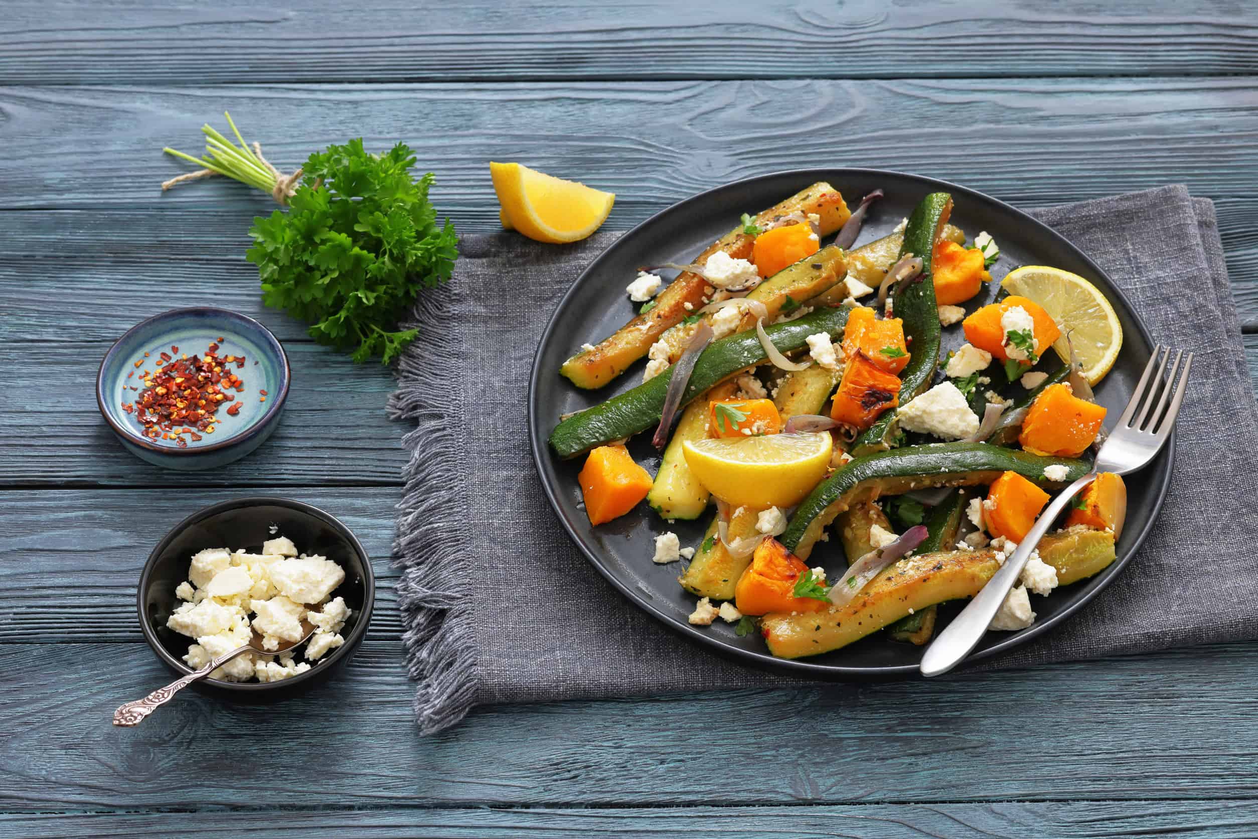 Plate of roasted vegetables with feta cheese and lemon wedges on a blue table, inspired by Vegetarian Dishes in Tunisia, with fresh herbs and seasonings nearby.