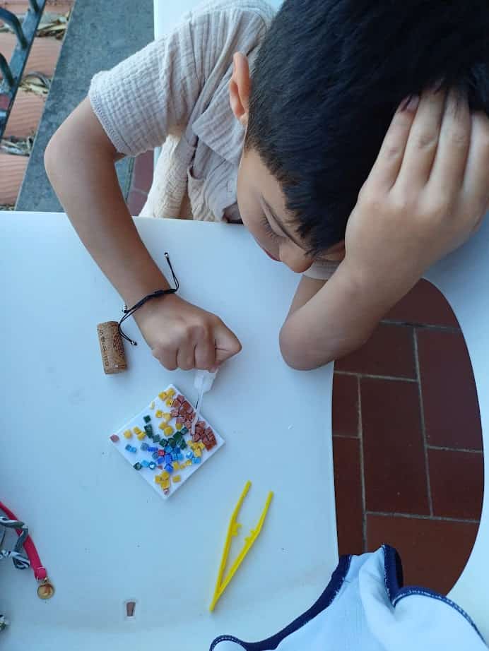 A child arranges colorful beads on a white square, with yellow tweezers and a cork nearby—much like a creative moment you might enjoy during 4 days in Castel Gandolfo.