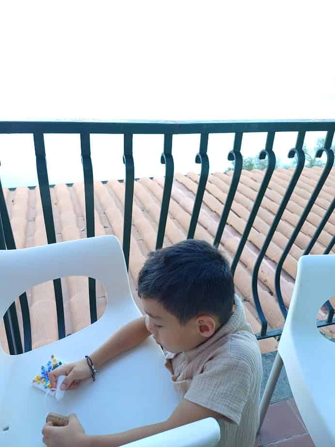 A young boy sits on a white chair by a railing, playing with colorful beads on a table, imagining he's spending 4 days in Castel Gandolfo.