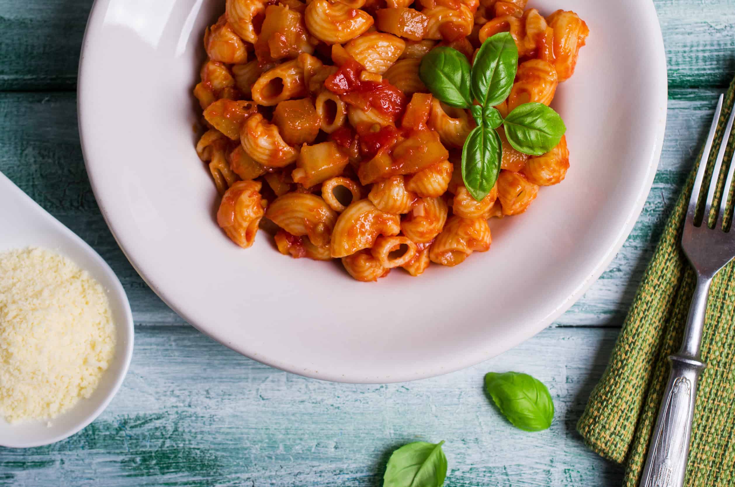 A plate of pasta with tomato sauce and basil, inspired by Vegetarian Dishes in Tunisia, sits next to a fork, napkin, and grated cheese on a rustic table.