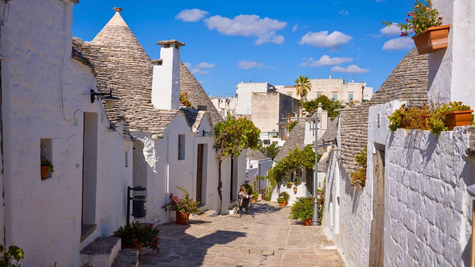 A sunny stone street with white cone-roofed houses, plants, and blue sky in Alberobello, Italy.