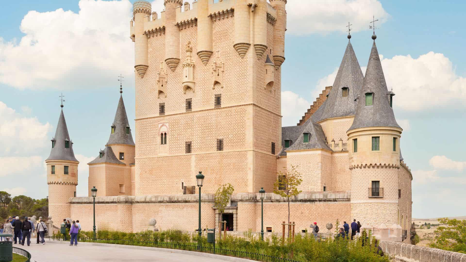 Historic stone castle with round towers and tall spires under a blue sky, visitors standing nearby.