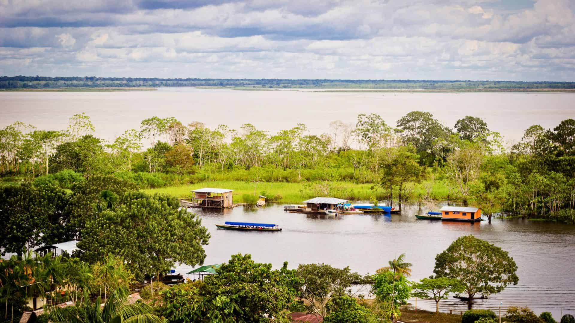River with boats and floating houses surrounded by lush green trees under a cloudy sky.