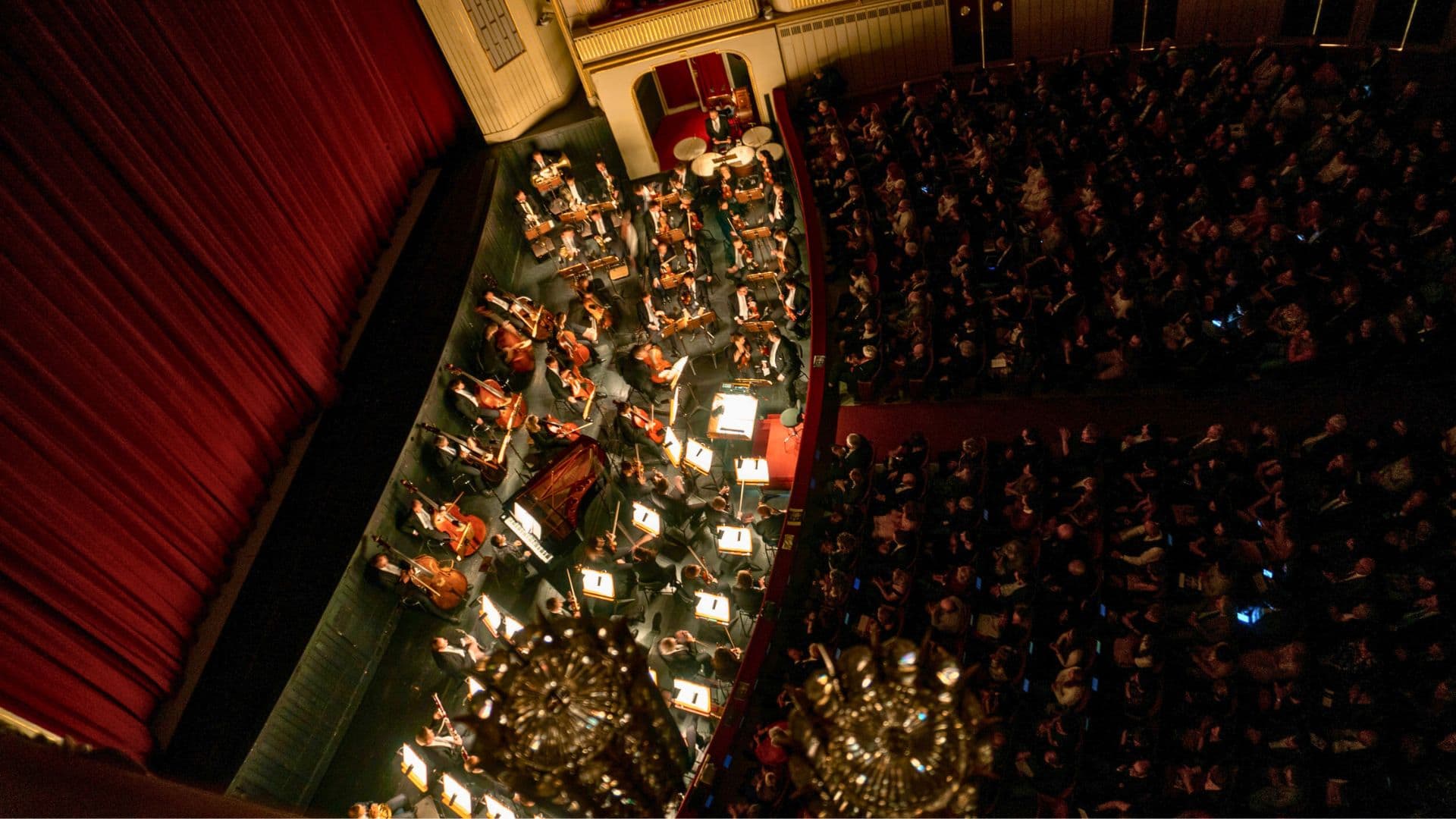 Aerial view of an orchestra performing on stage in a theater, with a large audience seated nearby.