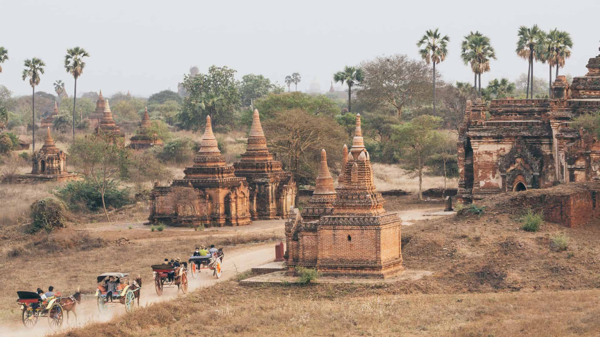 Horse-drawn carriages pass ancient temples and pagodas surrounded by dry grass and palm trees.