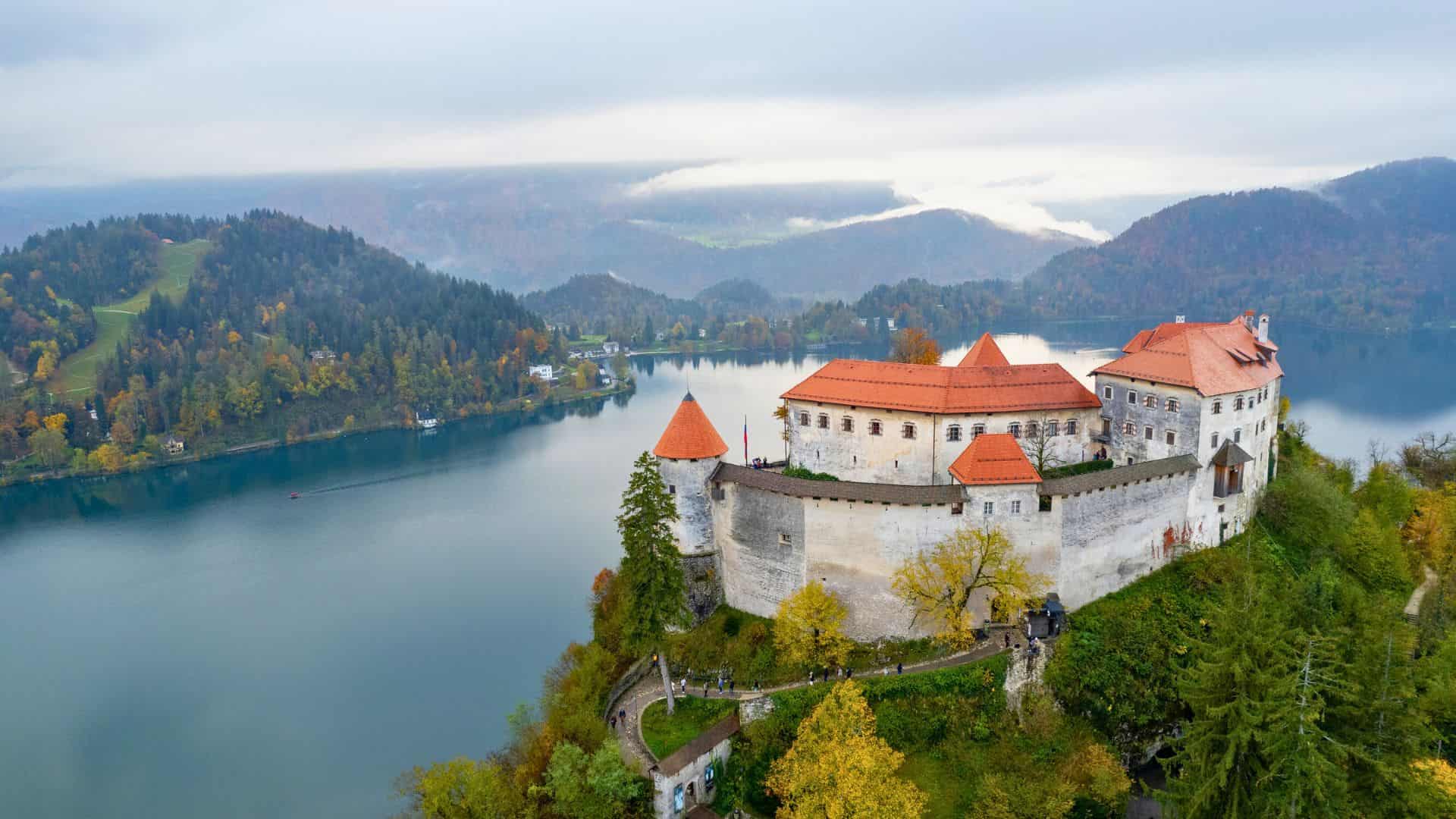 A castle with red roofs sits on a hill above a lake, surrounded by autumn trees and distant mountains.