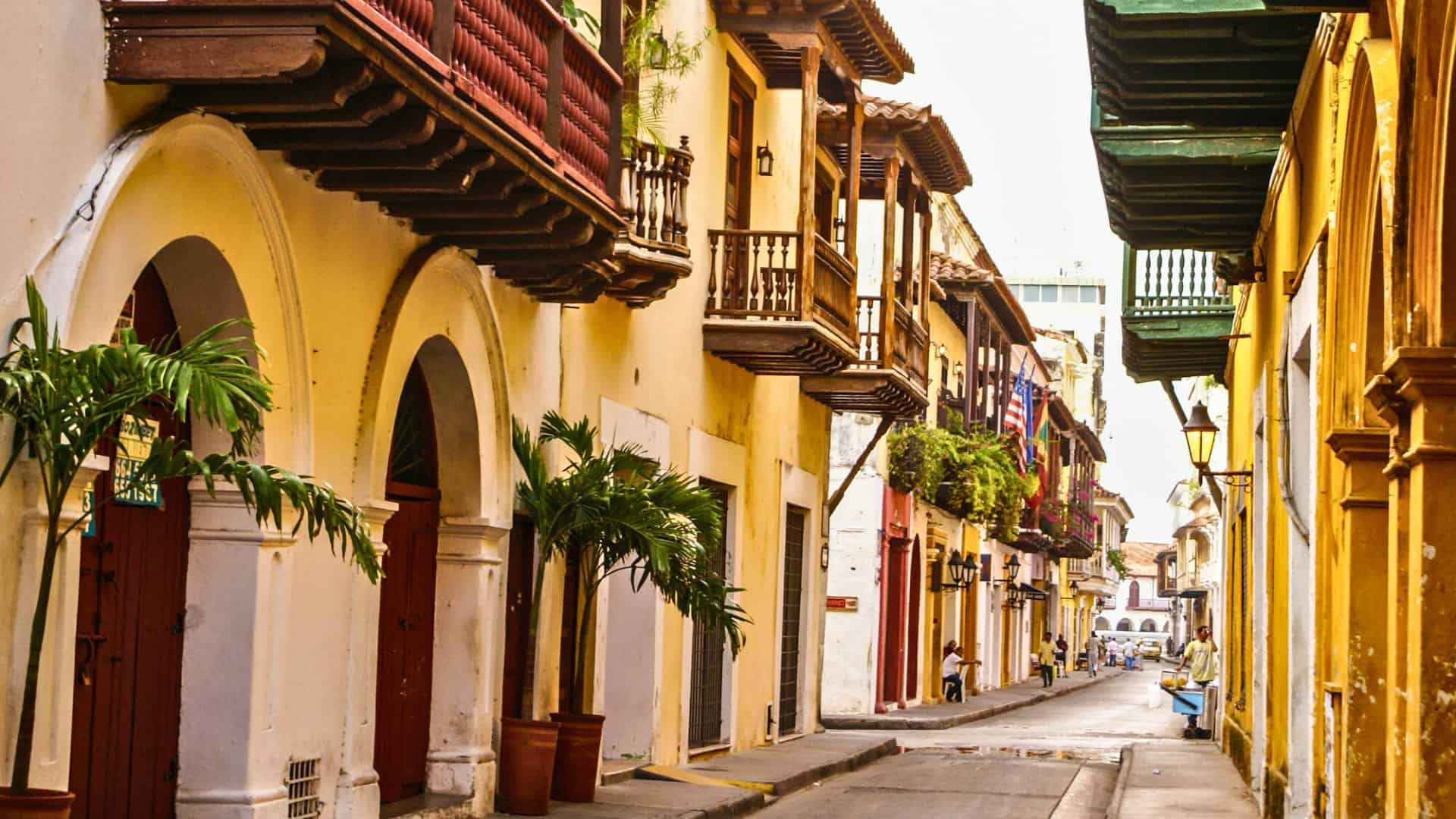 Colorful, narrow street lined with colonial buildings and balconies, with potted plants and a few people walking.