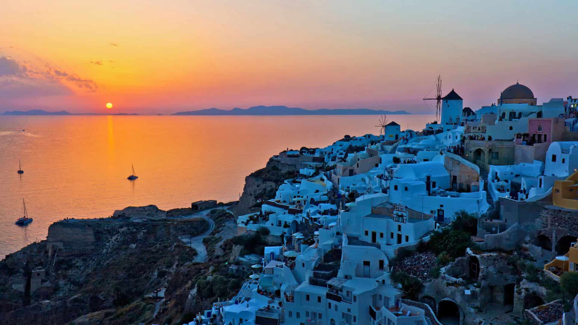 White buildings of Santorini overlook the sea at sunset, with boats on the water and a windmill in view.