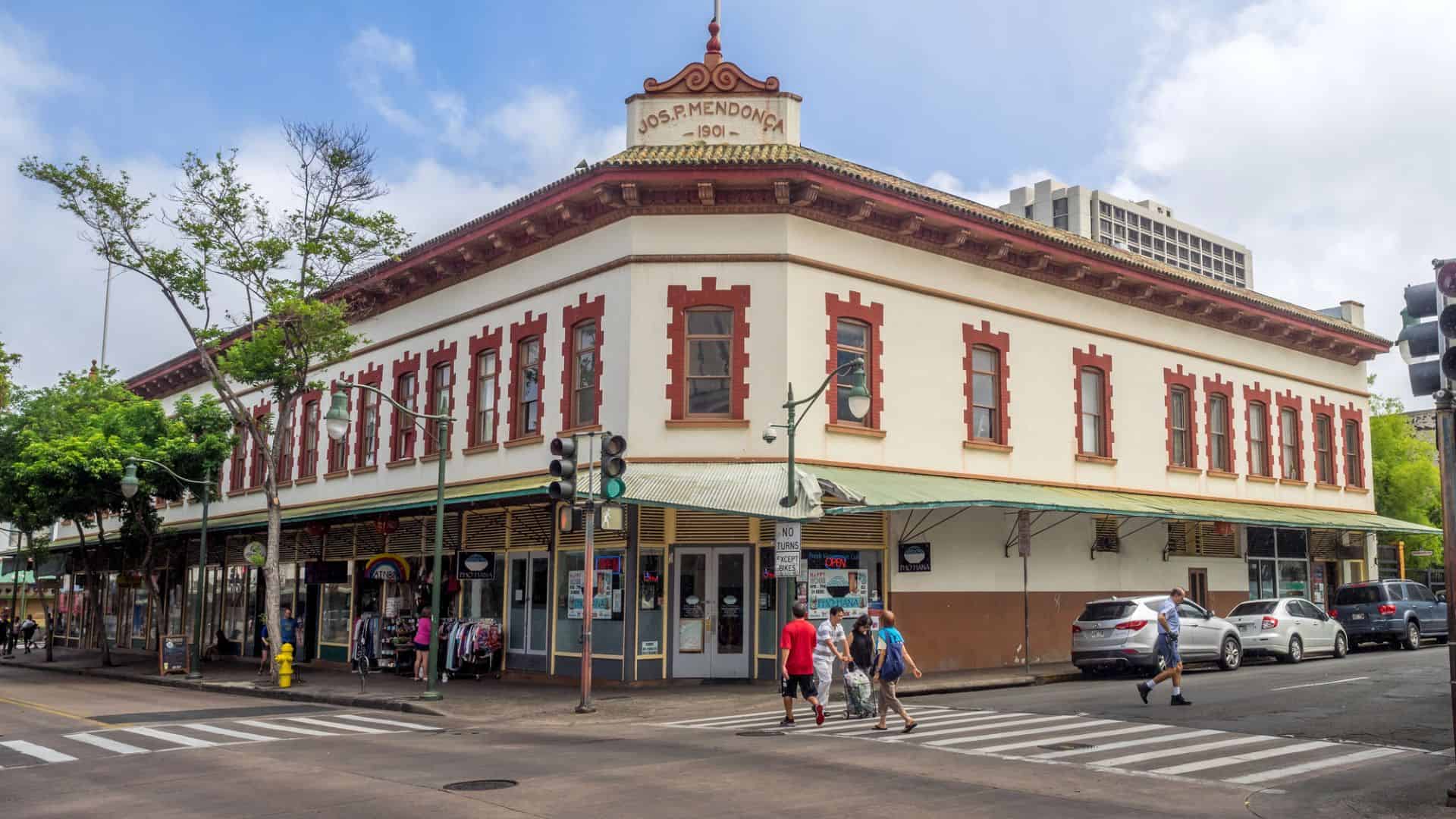 People cross the street by a historic two-story corner building with red trim and shops on the ground floor.