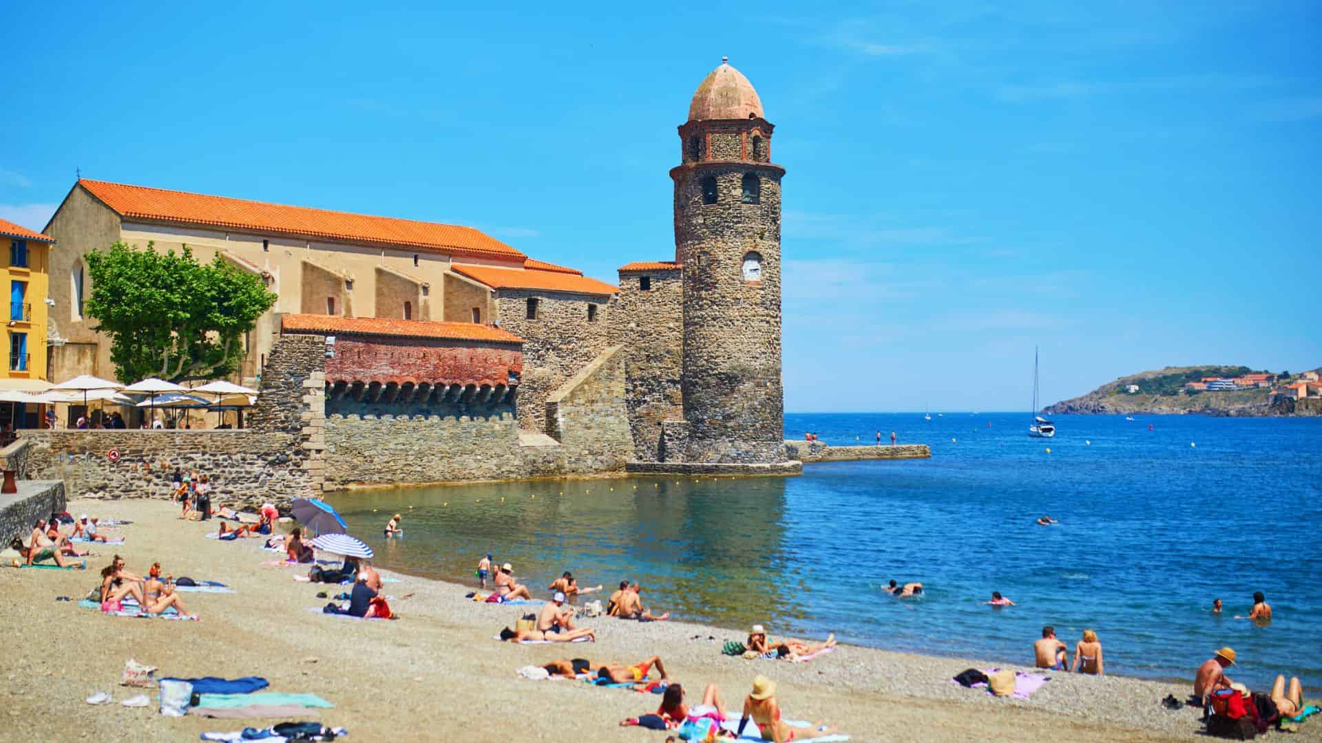 People relax and swim on a sunny beach near a historic stone church and tower by the sea.