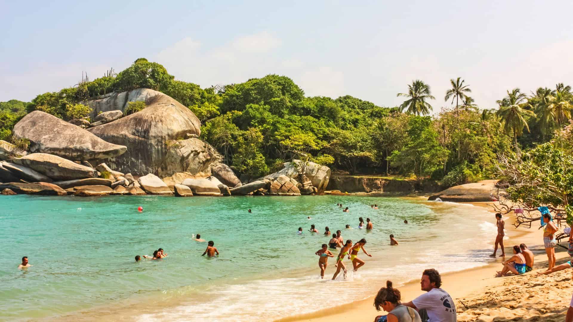People swimming and relaxing on a sandy beach with turquoise water, rocks, and lush green trees in the background.