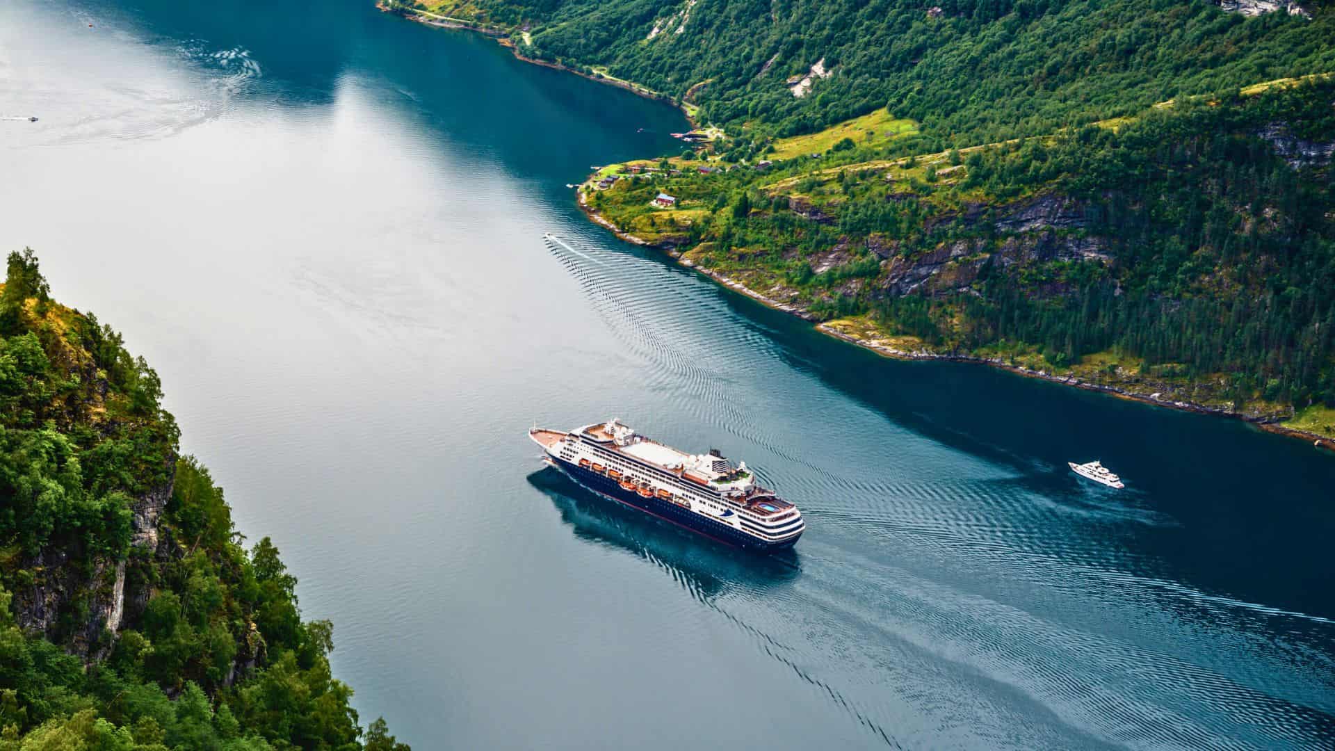 A cruise ship sails through a narrow, scenic fjord surrounded by green hills and rocky cliffs.