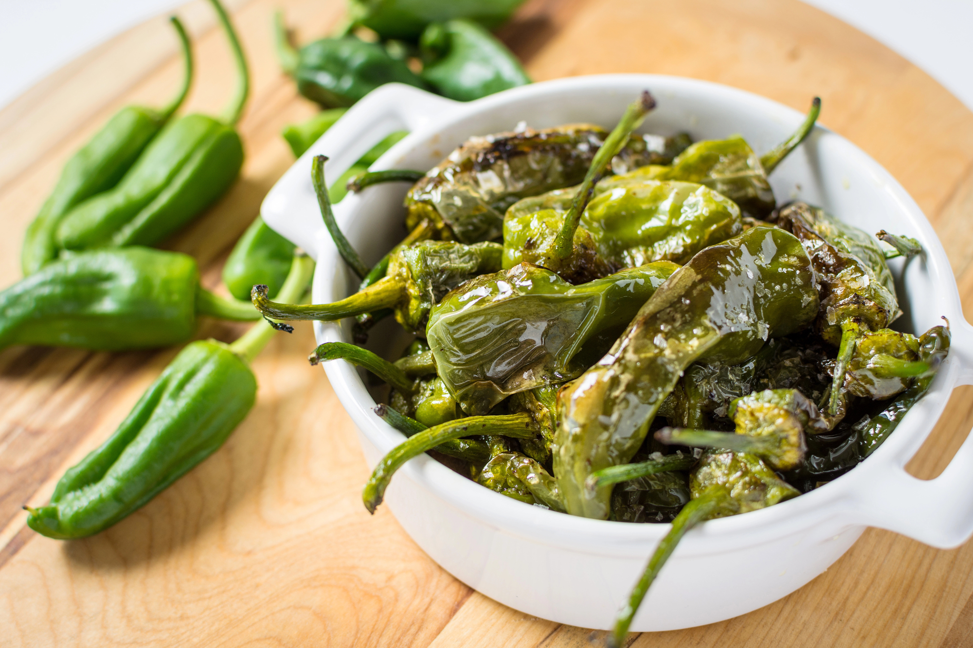 A white bowl filled with blistered, roasted green peppers&mdash;a classic vegetarian food from Spain&mdash;sits on a wooden cutting board with fresh peppers nearby.