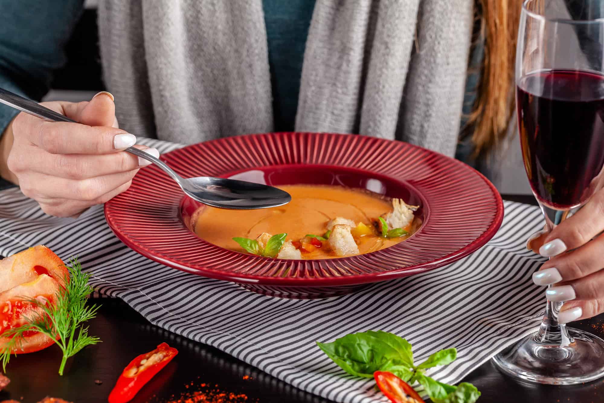 A person holds a spoon over a bowl of soup&mdash;one of the comforting vegetarian dishes Spain offers&mdash;with a glass of red wine on a striped napkin.