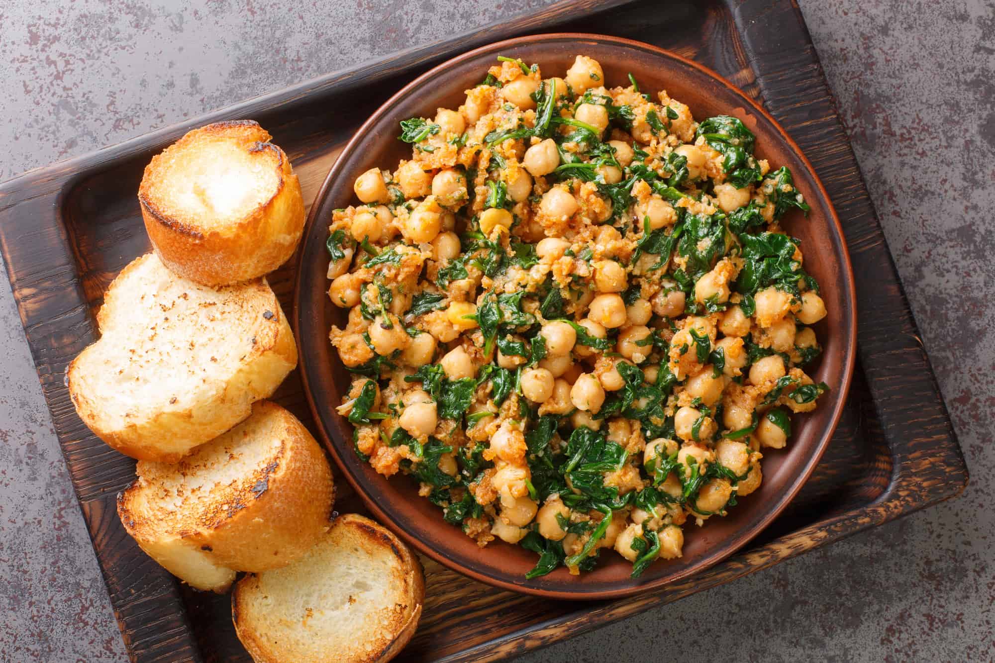 A bowl of saut&eacute;ed chickpeas with spinach, served with toasted bread slices on a wooden tray&mdash;a delicious example of vegetarian food often featured in Spain's top 10 favorite dishes.