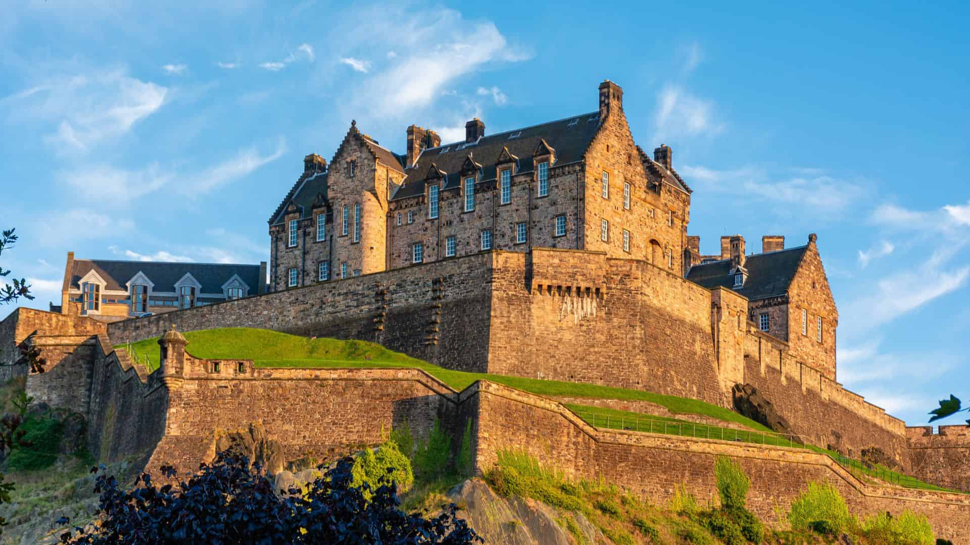 A historic stone castle sits atop a grassy hill under a blue sky with wispy clouds.