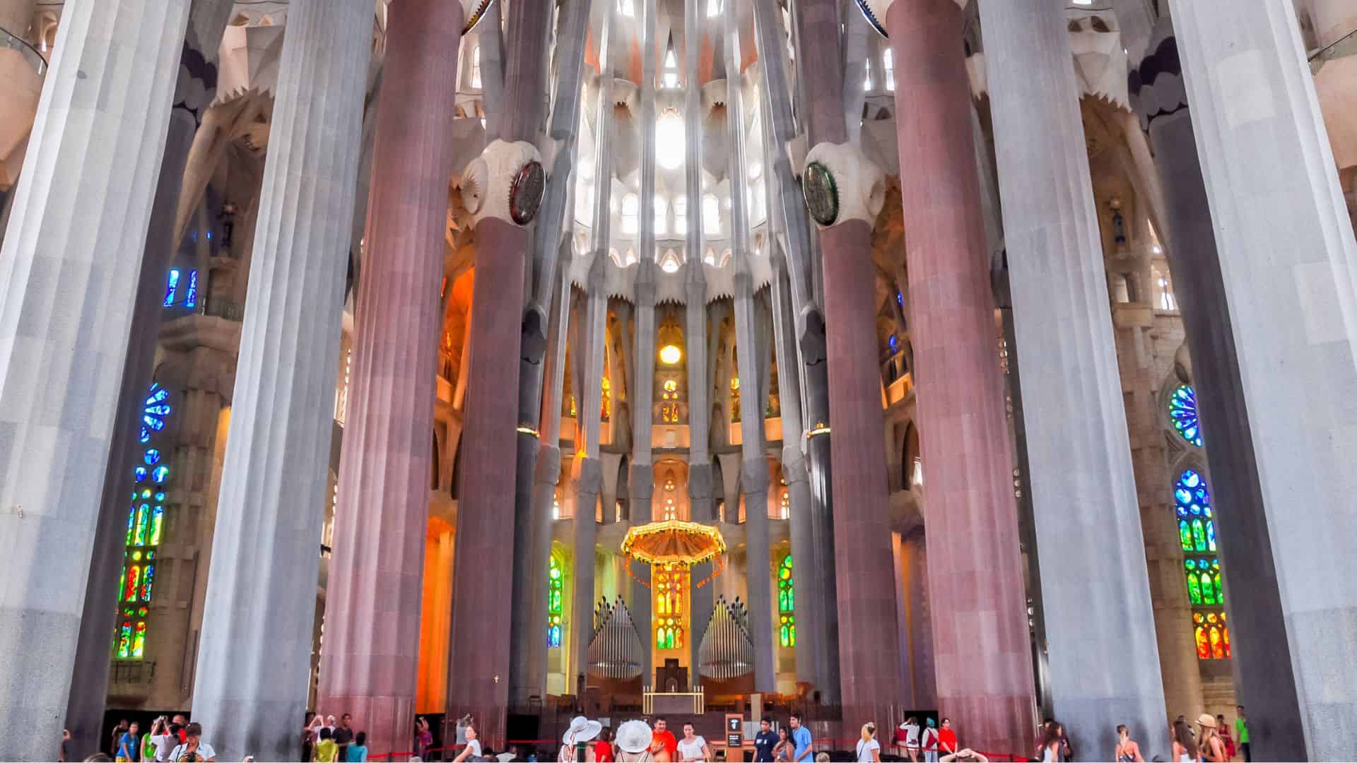Interior of Sagrada Familia with colorful stained glass, tall columns, and visitors walking inside.