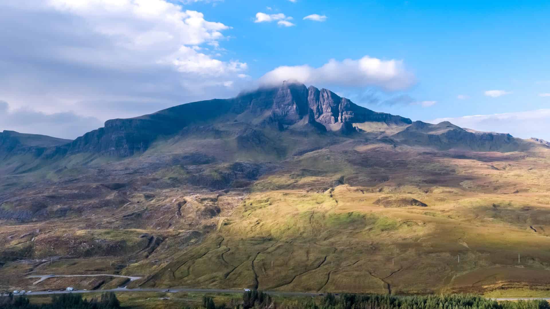 A scenic mountain landscape under a blue sky with clouds, green hills and a few roads in the foreground.