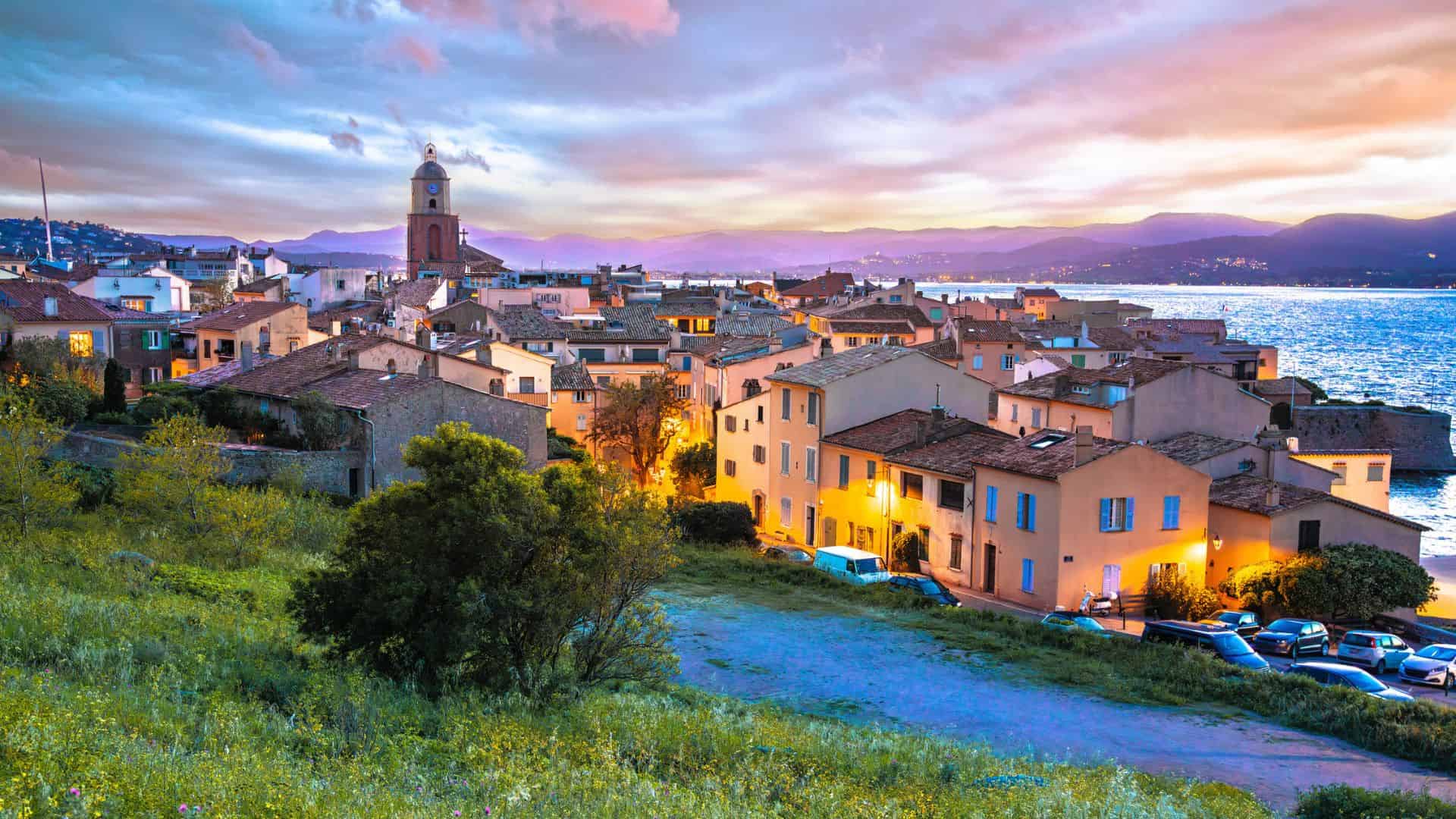 Colorful coastal town at sunset with lit buildings, a church tower, and mountains in the background.