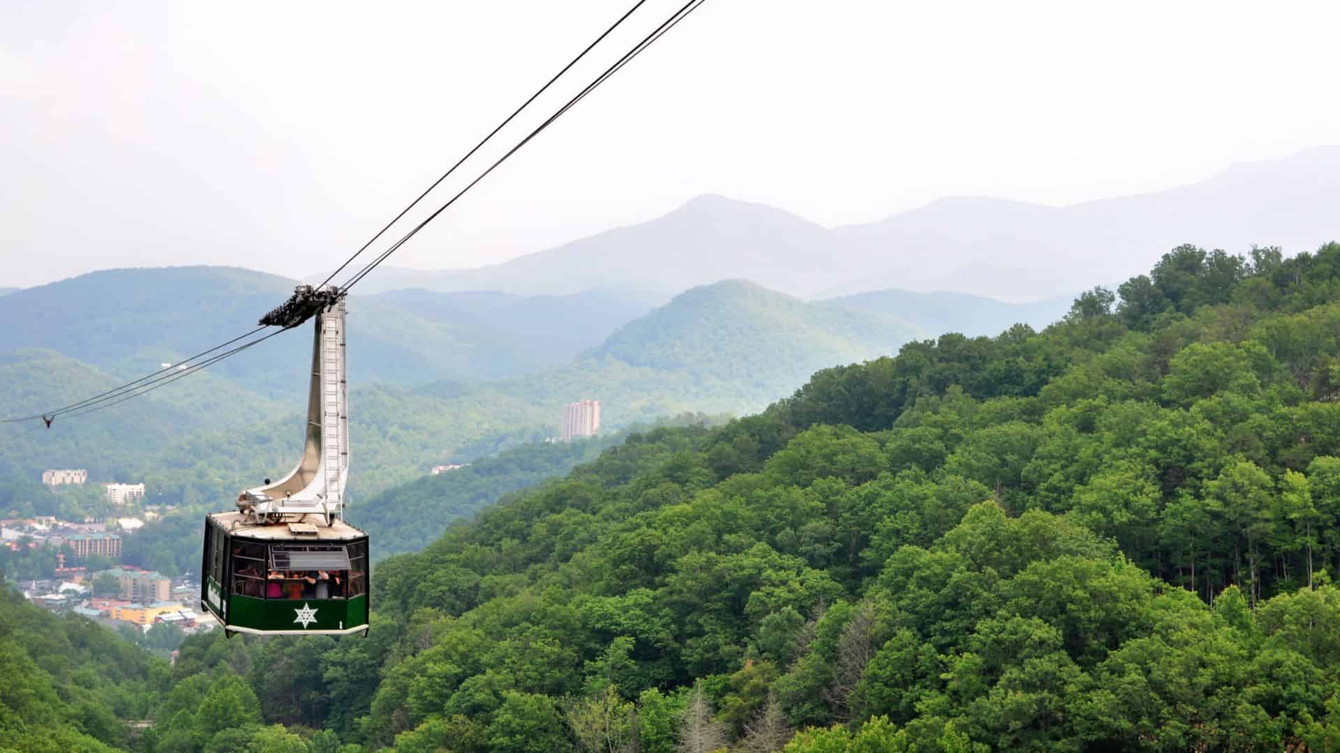 Green aerial tramway car travels above a lush, forested mountain landscape under a hazy sky.