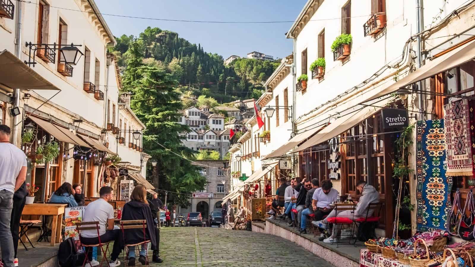 People sit at outdoor cafes along a cobblestone street lined with white buildings and colorful rugs.