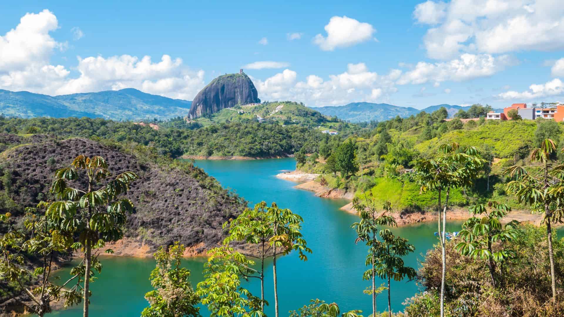 Large rock formation rises above lush green hills and blue lake under a bright sky with scattered clouds.