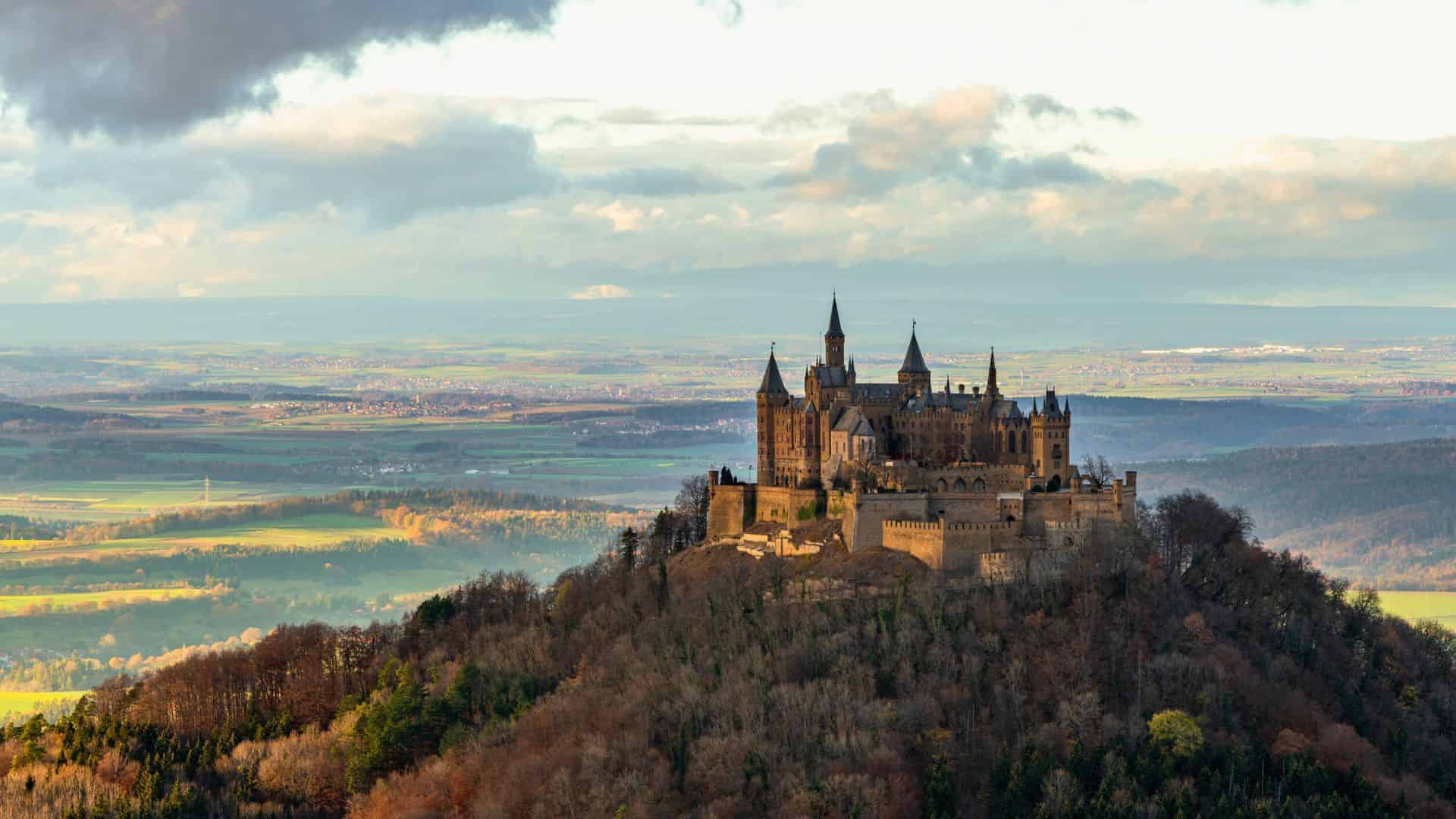 Castle atop a forested hill with sprawling fields and a cloudy sky in the background.