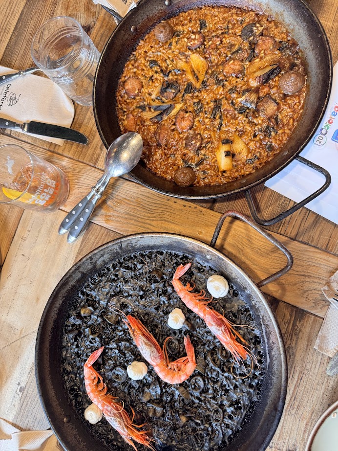 Two pans of Spanish rice dishes: one with shrimp and squid ink, the other showcasing Spanish vegetarian cuisine with chorizo-style veggie sausage and vegetables on a wooden table.