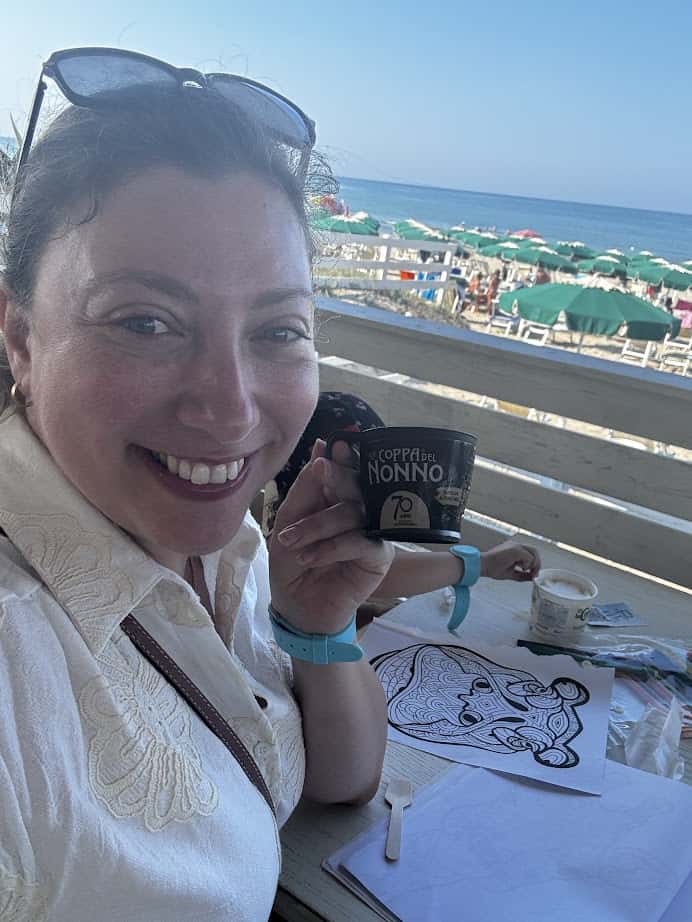 Smiling woman at a beachside caf&eacute; holds a cup, with coloring pages and the sea in the background&mdash;capturing the relaxed vibe of the best beach resorts in Italy for families.