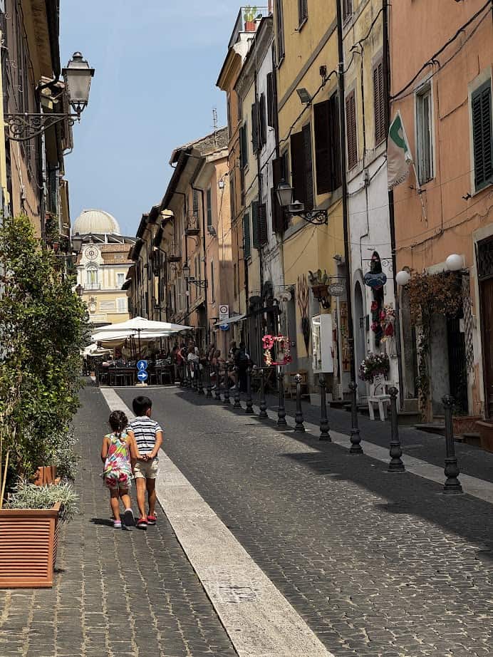 Two children walk down a cobblestone street lined with colorful buildings and outdoor cafes on a sunny day, capturing the charm you might experience during 4 days in Castel Gandolfo.