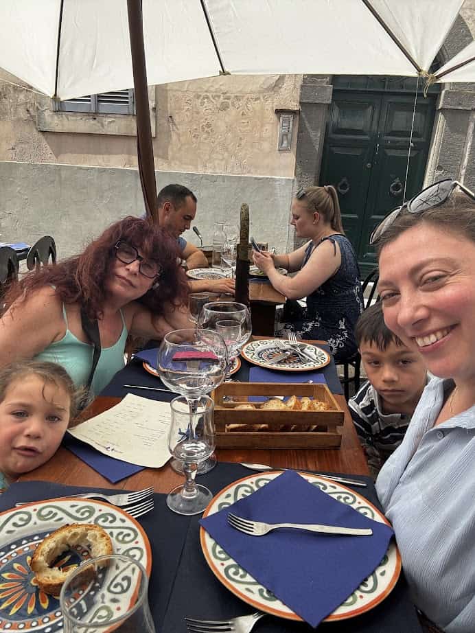 Four people sitting at an outdoor restaurant table with food and drinks, smiling under a white umbrella, sharing stories about the best day trips from Rome for families.