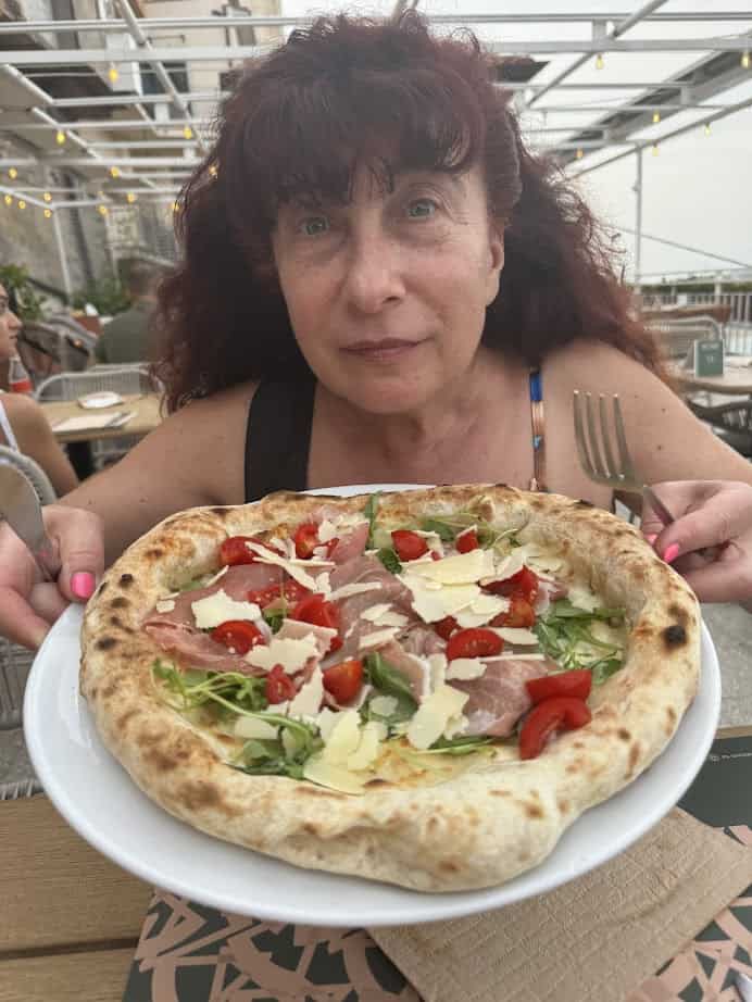 Savoring 4 days in Castel Gandolfo, a woman holds a plate with fresh pizza topped with prosciutto, arugula, cherry tomatoes, and cheese flakes.