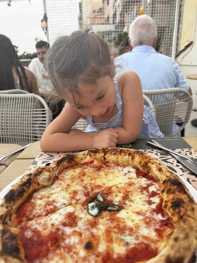 A young girl eagerly looks at a freshly baked margherita pizza on a table at an outdoor restaurant, enjoying one of the highlights of her 4 days in Castel Gandolfo.