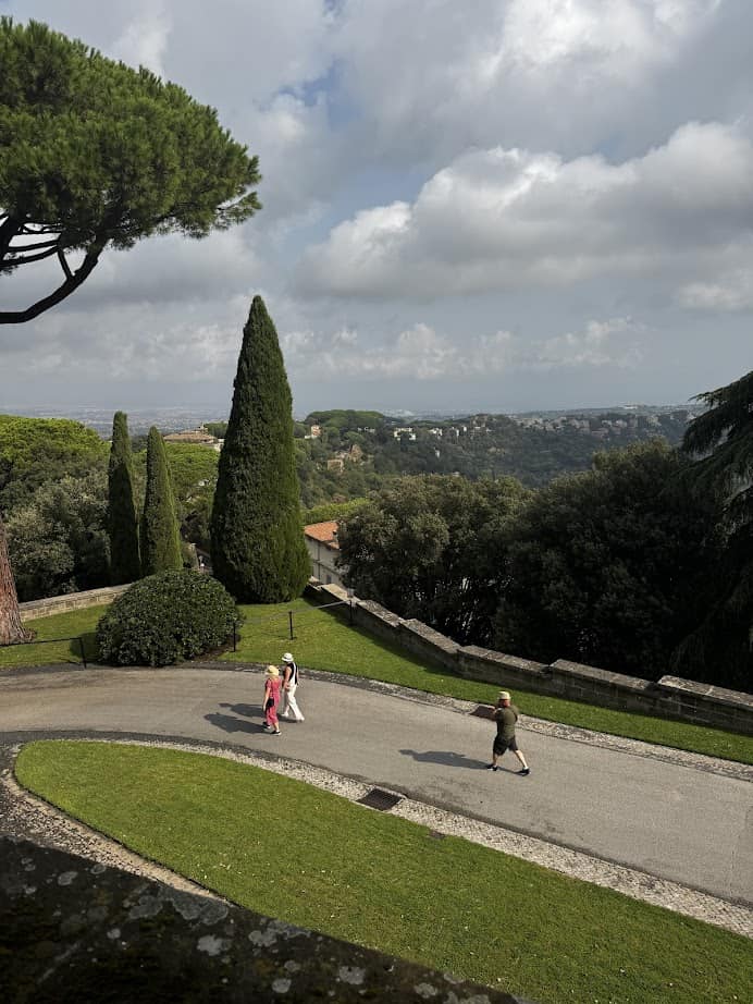 Three people walk on a curved path in a lush, hilly park with trees and distant buildings under a partly cloudy sky, enjoying their 4 days in Castel Gandolfo.