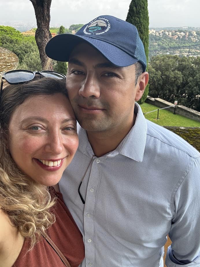 A smiling woman and a man in a blue cap pose together outdoors with a scenic landscape in the background, capturing memories from their 4 days in Castel Gandolfo.
