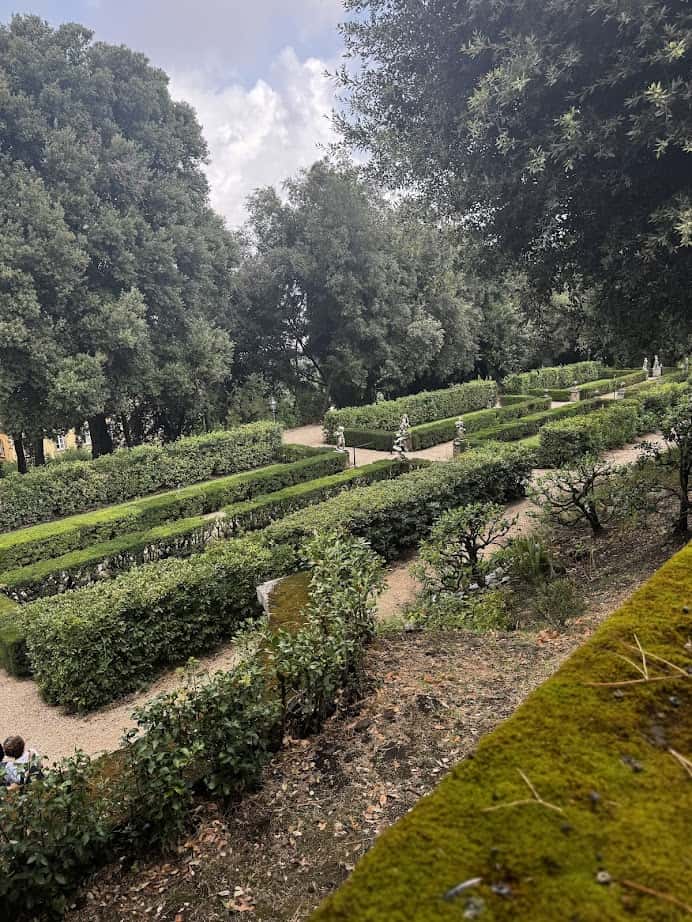 Elegant formal garden with neatly trimmed hedges, gravel paths, and statues—perfect for exploring during 4 days in Castel Gandolfo—set amid tall trees under a cloudy sky.