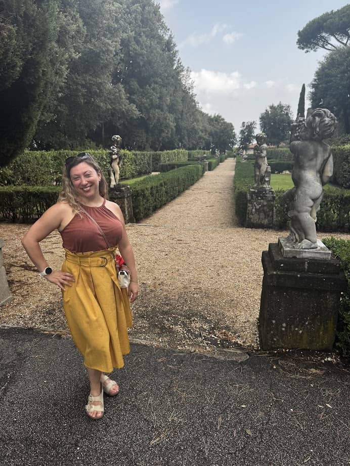 Smiling woman in a yellow skirt stands in a formal garden with statues and manicured hedges, enjoying her 4 days in Castel Gandolfo.