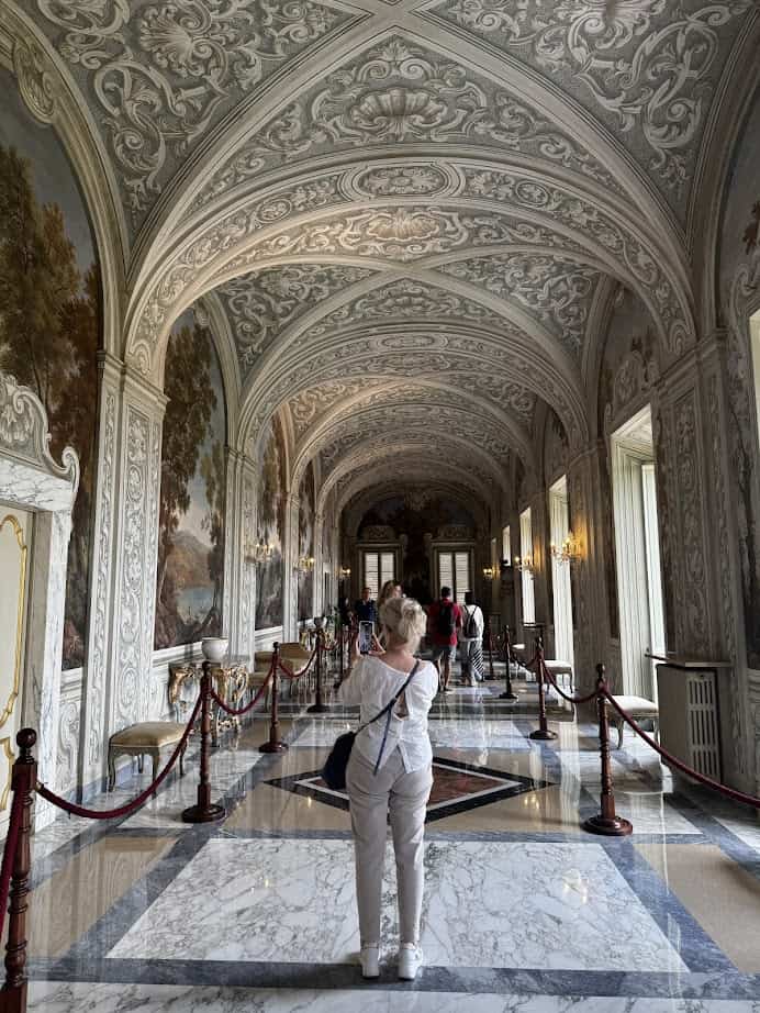 A woman stands in an ornate, marble hallway with painted ceilings and walls, capturing a memory during her 4 days in Castel Gandolfo.