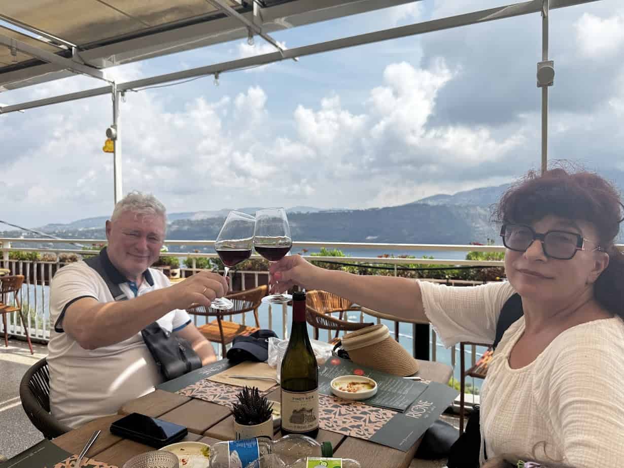 Two people toast with wine glasses at an outdoor restaurant, enjoying a scenic mountain and water view—an unforgettable moment during 4 days in Castel Gandolfo.