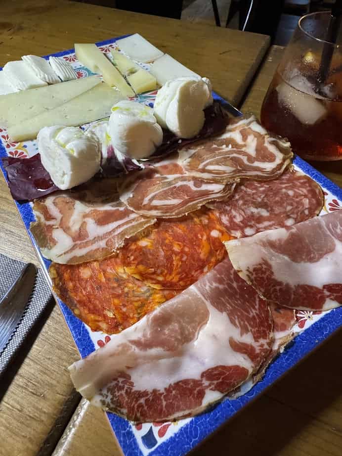 A platter with assorted sliced meats, cheeses, and mozzarella balls captures the flavors of 4 days in Castel Gandolfo, served next to a red drink with ice.