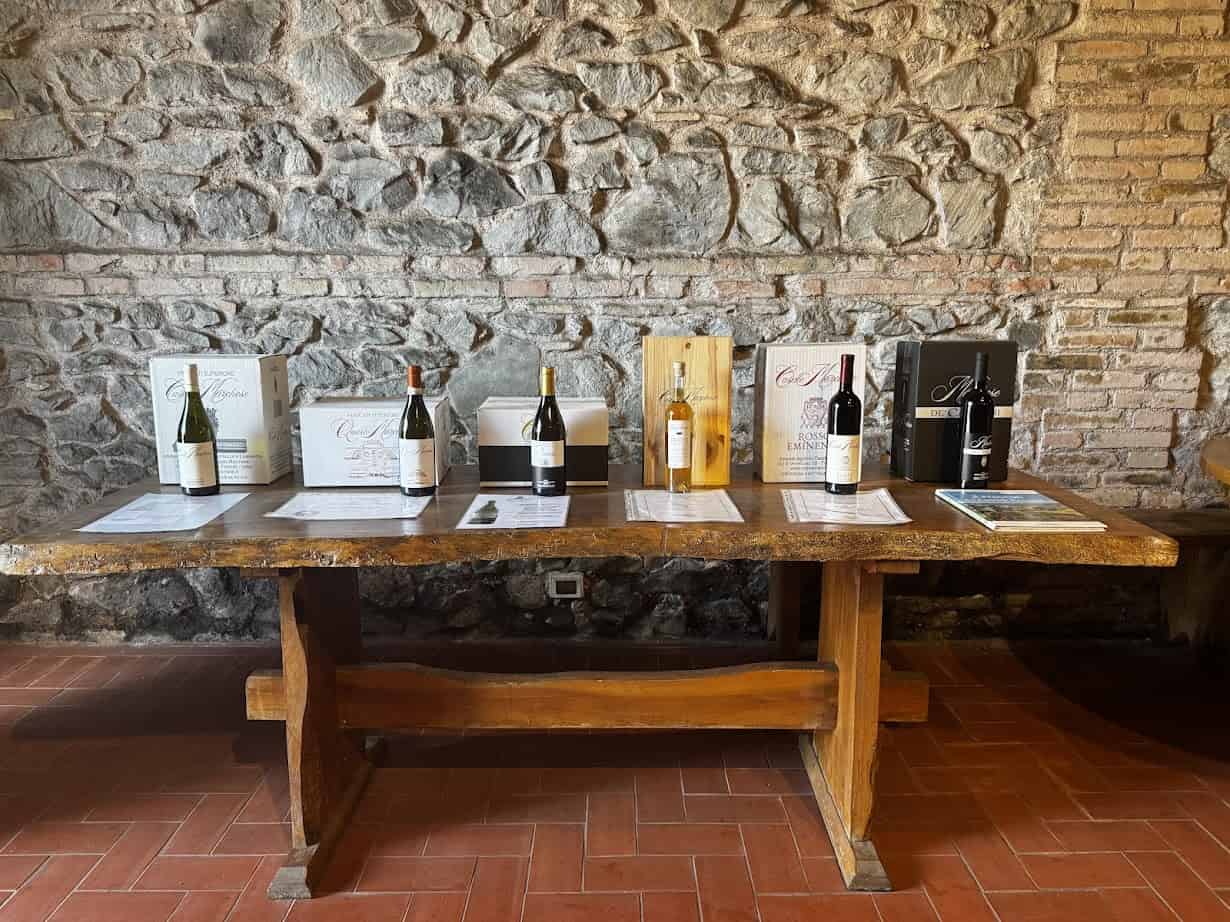 Bottles of wine with display cards arranged on a rustic wooden table against a stone wall evoke the charm of spending 4 days in Castel Gandolfo.