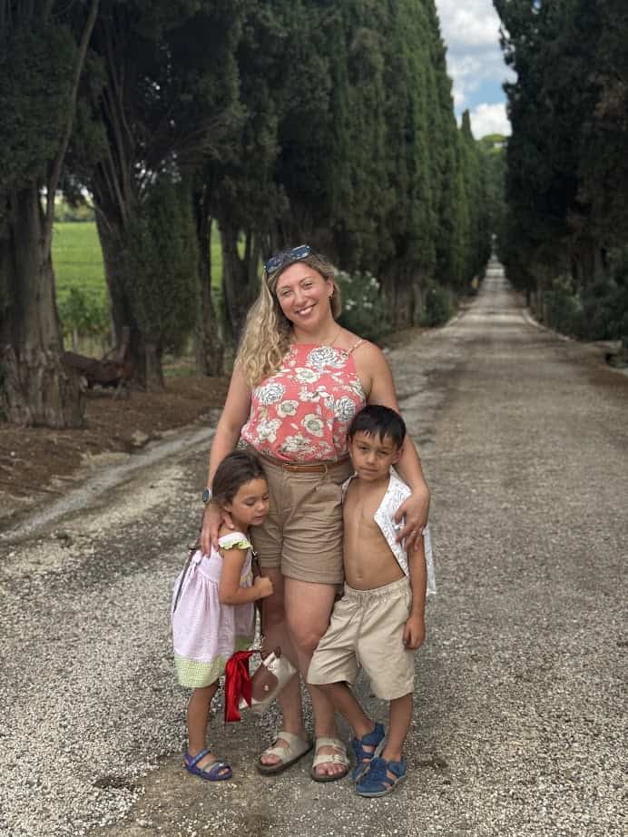 A smiling woman stands on a tree-lined path with two young children hugging her on each side, enjoying precious moments together during their 4 days in Castel Gandolfo.